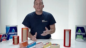 Professional painter in navy blue shirt demonstrating paint products while seated at a white table with various Armstead paint cans and color samples.
