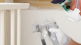 Person wearing white protective clothing using a spray gun to paint white trim or molding indoors.