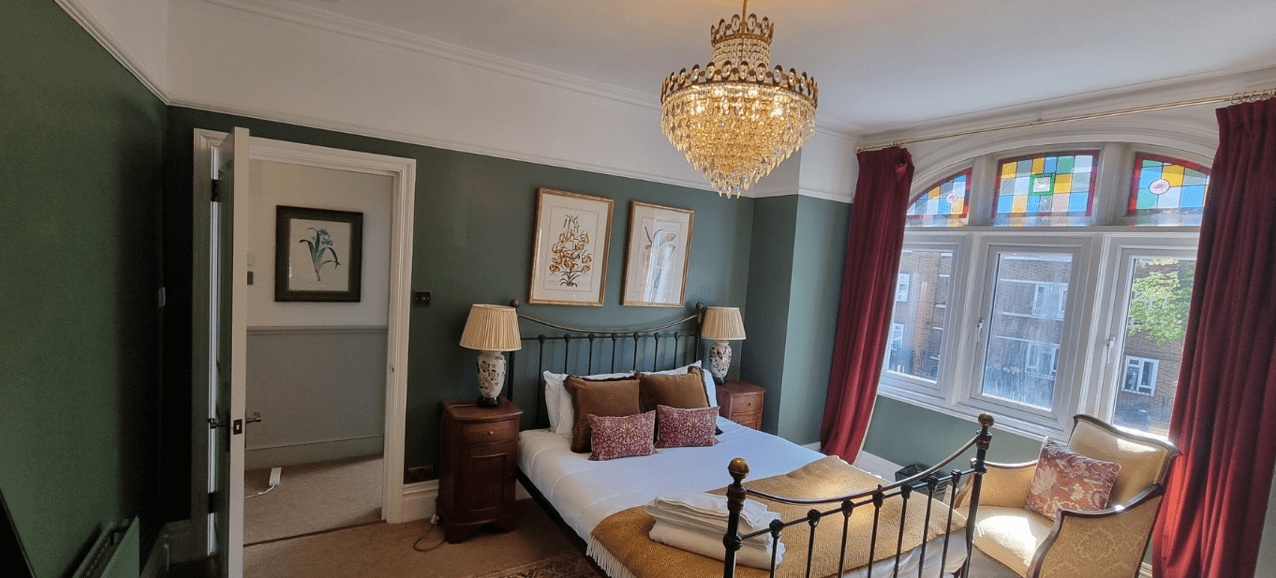 Elegant Victorian-style bedroom with sage green walls, crystal chandelier, black metal bed frame, stained glass windows, and burgundy curtains.