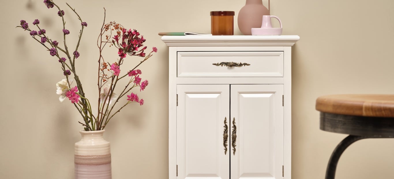 Eine restaurierte Kommode vor einer Wand, daneben eine Vase mit Blumen und ein Hocker