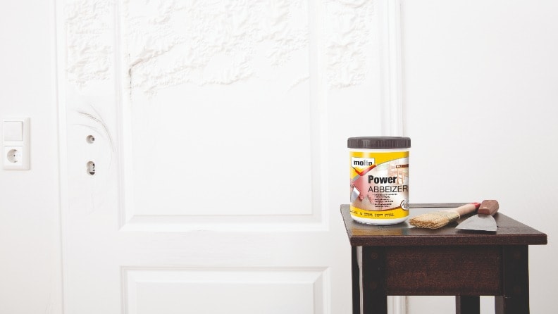 Weiße Tür, an der sich nach dem Auftragen eines Abbeizers die Farbschicht ablöst, davor ein Hocker mit Werkzeug und einer Dose Power Abbeizer von Molto