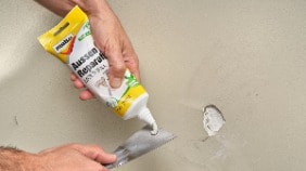 Spachtelmasse wird auf einen Spachtel gedrückt vor einer Wand im Außenbereich mit einem Loch im Putz