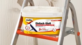 Zimmerwand vor dem Renovieren mit Löchern, davor eine Frau mit Farbroller in der Hand und daneben eine Trittleiter