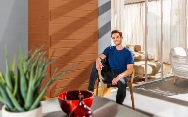 Mann sitzt auf einer Terrasse vor einer in Rehbraun gestrichenen Fassade 