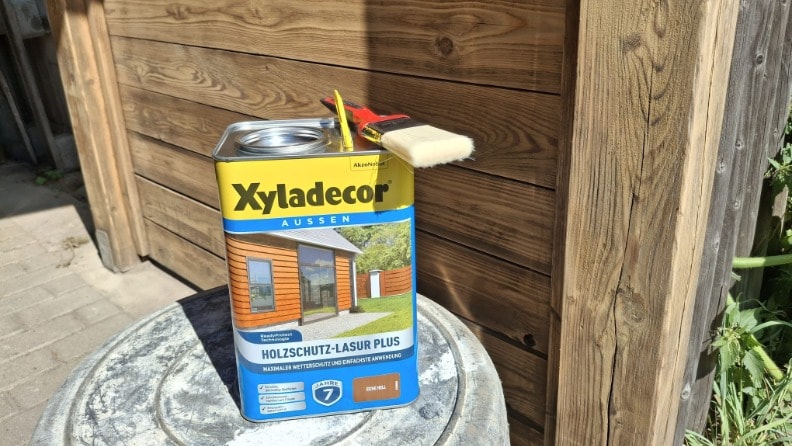 Eine Dose Holzschutz-Lasur Plus von Xyladecor mit einem Flachpinsel vor einem Carport