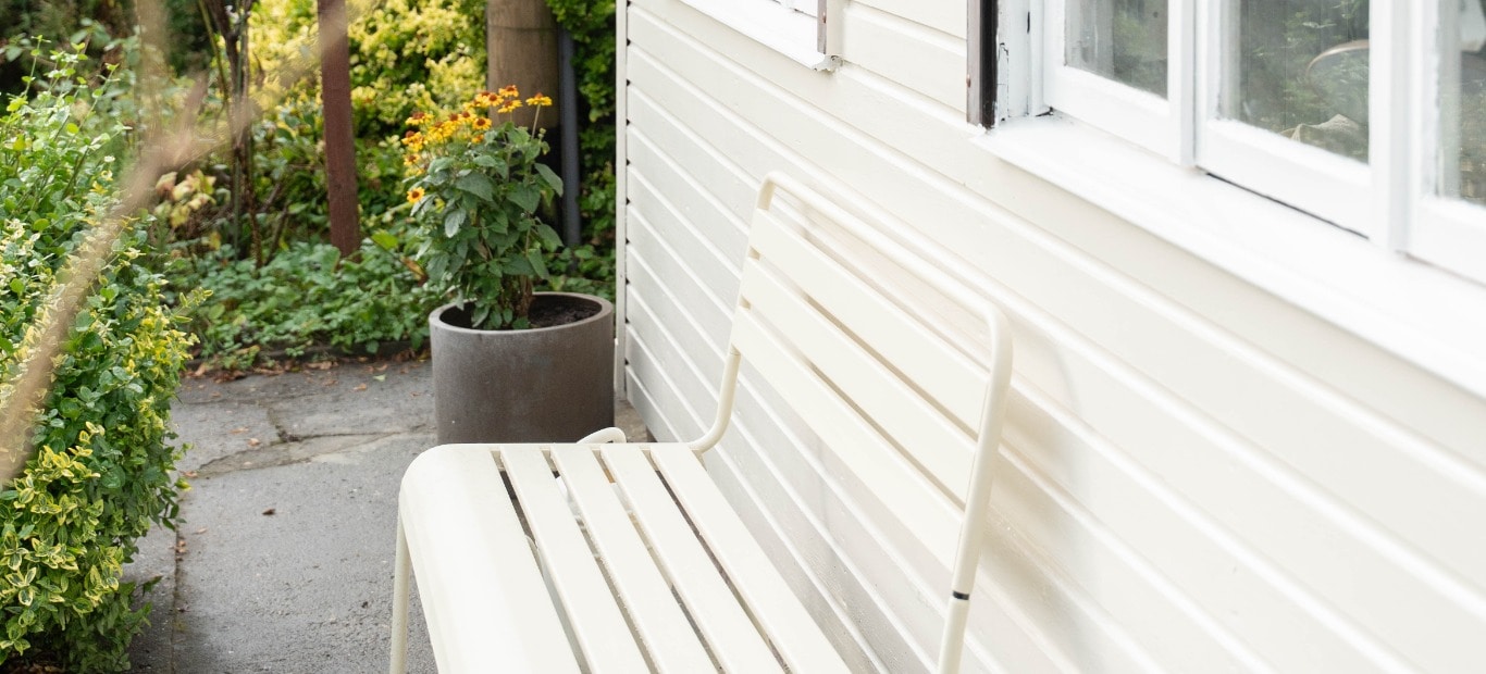 In Kieselweiß gestrichenes Gartenhaus aus Holz mit Sitzebank umgeben von einem Steinweg und Pflanzen