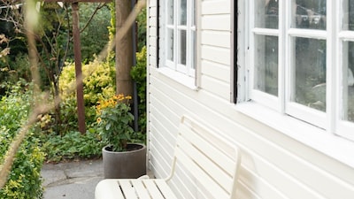 Mit Conslan Kieselweiß gestrichenes Gartenhaus aus Holz mit Sitzebank im Garten umgeben von einem Steinweg undPflanzen