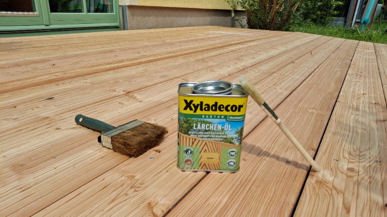 Eine mit Lärchen-Öl von Xyladecor frisch gestrichene Holzterrasse mit Pinseln und einer Dose darauf 